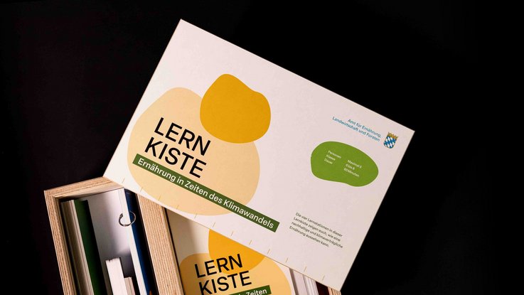 VISUELL Kommunikationsdesign: Konzeption und Realisierung einer pädagogischen Lernkiste zum Thema: Ernährung in Zeiten des Klimawandels für Klasse 6 –8. VISUELL Szenografie: Pädagogische Lernkiste: Holzkiste mit Deckel mit grün und gelben organischen Elemente- Aufschrift: Lernkiste, Ernährung in Zeiten des Klimawandels