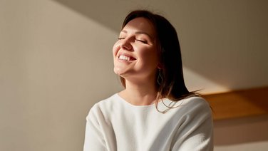 VISUELL Kommunikationsdesign: Corporate Design: Eine junge Frau mit dunklen Haaren, geschlossenen Augen und strahlendem Lächeln streckt ihr Gesicht der Sonne entgegen