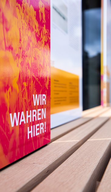 VISUELL Szenografie: Informationseinheiten für die sieben UNESCO-Welterbestätten in Baden-Württemberg in Zusammenarbeit mit dem Ministerium für Landesentwicklung und Wohnen Baden-Württemberg VISUELL Szenografie: Wahre Wunderbänke: Nahaufnahme einer Sitzbank mit Informationstafel mit plakatives Bildmotiv in magenta und orange mit der Aufschrift WIR WAHREN HIER!