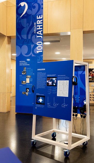 VISUELL Szenografie: Jubiläumsausstellung: Im Vordergrund steht ein Rollmöbel mit integrierten Displays, Hörstation, grafischen Abbildungen und Text; im Hintergrund hängt ein großflächiger Banner