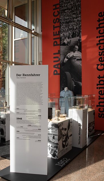 VISUELL Szenografie: Jubiläumsausstellung: Vom Rennfahrer zum Verleger, 75 Jahre Verlagsgeschichte VISUELL Szenografie: Ausstellungsrundgang: Bereich "Der Rennfahrer" - Stoffbanner Aufschrift: Paul Pietsch schreibt Geschichte