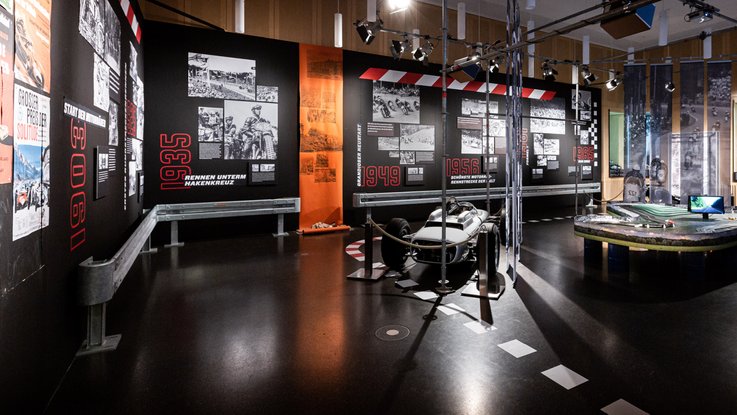 VISUELL Szenografie: Mythos Solitude: Oldtimer und Carrera-Bahn im Vordergrund und schwarze Informationstafeln im Hintergrund mit schwarz weiß Bildern umrandet von Leitplanken