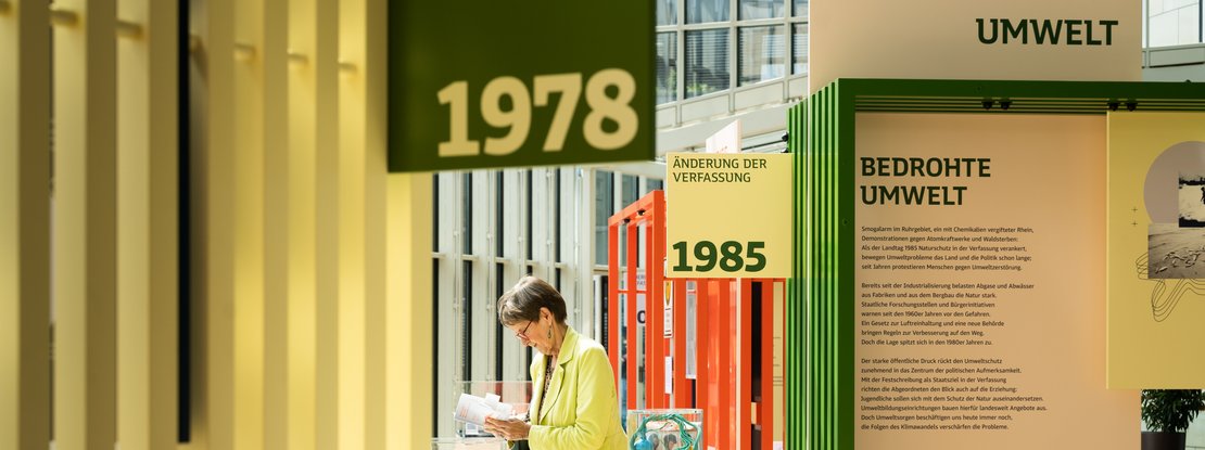 VISUELL Szenografie: Wanderausstellung „Jubiläum. 75 Jahre Verfassung NRW“ für die Stiftung Haus der Geschichte Nordrhein-Westfalen VISUELL Szenografie: Wanderausstellung: Nahaufnahme der Themenmodule Umwelt und Verfassungsänderung. Im Vordergrund gelbe Lamellenstruktur mit den Jahreszahlen 1978 und 1985, eine Besucherin liest in einem Buch.