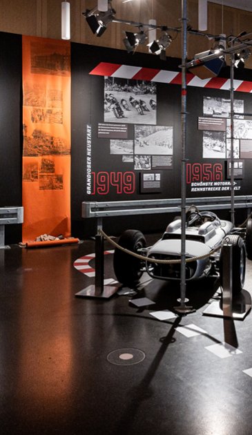 VISUELL Szenografie: Mythos Solitude: Oldtimer im Vordergrund und schwarze Informationstafeln im Hintergrund mit schwarz weiß Bildern- Aufschrift in Rot: Grandioser Neustart 1949 und Schönste Motorradrennstrecke der Welt 1956 