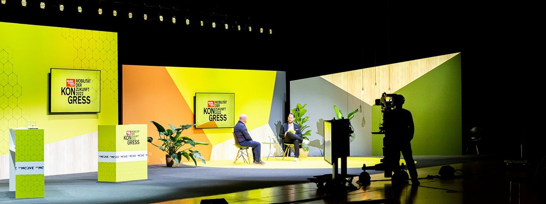 VISUELL Szenografie: Bühnenbild und Gesamtinszenierung vom AMS Kongress, Auto Motor und Sport, Motor Presse Stuttgart VISUELL Szenografie: Zwei Personen unterhalten sich vor einer gelb/orangenen Wand während sie gefilmt werden