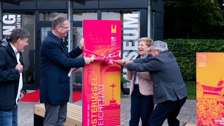 VISUELL Szenografie: Informationseinheiten für die sieben UNESCO-Welterbestätten in Baden-Württemberg in Zusammenarbeit mit dem Ministerium für Landesentwicklung und Wohnen Baden-Württemberg VISUELL Szenografie: Wahre Wunderbänke: Einweihung der Informationseinheit, vier Personen ziehen an einer Schleife, die um eine Informationstafel gebunden ist