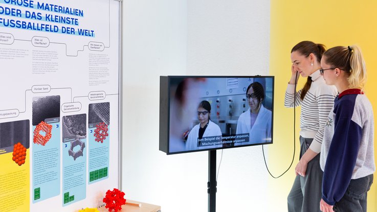 VISUELL Szenografie: Wissenschaftsausstellung „Was kann Katalyse?“ für den Sonderforschungsbereich SFB 1333 der Universität Stuttgart VISUELL Szenografie: Wissenschaftsausstellung: Zwei Besucherinnen stehen vor einer Bildschirmstele und verfolgen den abgespielten Film, eine von ihnen hält einen Handkopfhörer an ihr Ohr. Daneben befindet sich ein Plakat mit Informationen und mehrere Modelle von Molekülstrukturen von unterschiedlichen Materialien.