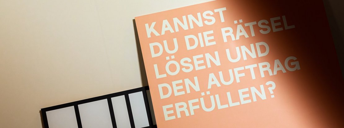 VISUELL Szenografie: 200 Jahre KOSMOS, Jubiläumsausstellung VISUELL Szenografie: Detailaufnahme orangene Texttafel – Aufschrift: Kannst du die Rätsel lösen und den Auftrag erfüllen?