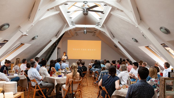 VISUELL Blogbeitrag: Das Event unterm Dach mit dem Thema „Taste the best“ – das Olivenöl-tasting. VISUELL Blogbeitrag: Event unterm Dach– das Olivenöl-tasting: Viele Leute sitzen an Tischen und hören einem Vortrag zu