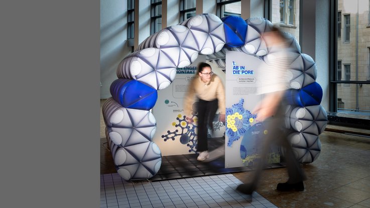 VISUELL Szenografie: Wissenschaftsausstellung „Was kann Katalyse?“ für den Sonderforschungsbereich SFB 1333 der Universität Stuttgart VISUELL Szenografie: Eine Besucherin steht in einem begehbaren, aufgeblasenen Modell einer Pore in der Wissenschaftsausstellung „Was kann Katalyse?“. Die Struktur des Modells besteht aus Molekülen und ist als Bogen aufgebaut. Im Inneren der Pore befindet sich mehrere Informationstafeln. Eine weitere Besucherin läuft im Vordergrund an dem Exponat vorbei.