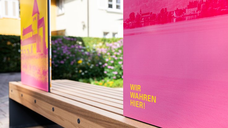 VISUELL Szenografie: Informationseinheiten für die sieben UNESCO-Welterbestätten in Baden-Württemberg in Zusammenarbeit mit dem Ministerium für Landesentwicklung und Wohnen Baden-Württemberg VISUELL Szenografie: Wahre Wunderbänke: Rückseite einer Sitzbank mit Informationstafeln in magenta und gelb mit der Aufschrift WIR WAHREN HIER!
