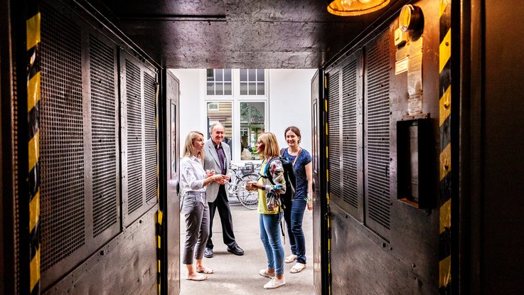 Titel: VISUELL Blogbeitrag: Das Event unterm Dach mit dem Thema „Taste the best“ – das Olivenöl-tasting. VISUELL Blogbeitrag: Event unterm Dach– das Olivenöl-tasting: Vier Personen stehen im Vorhof von VISUELL und unterhalten sich
