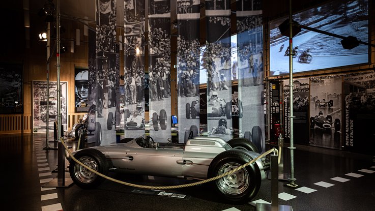 VISUELL Szenografie: Mythos Solitude: Oldtimer in der Mitte des Raumes und schwarz weiße Stoffbanner hängen von der Decke darunter