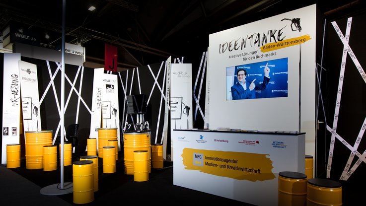 VISUELL Szenografie: Messestand für die MFG auf der Frankfurter Buchmesse VISUELL Szenografie: Messestand Ideentanke MFG: Gesamtüberblick über den Messestand mit gelben Sitztonne, typografischen Bannern und Screens
