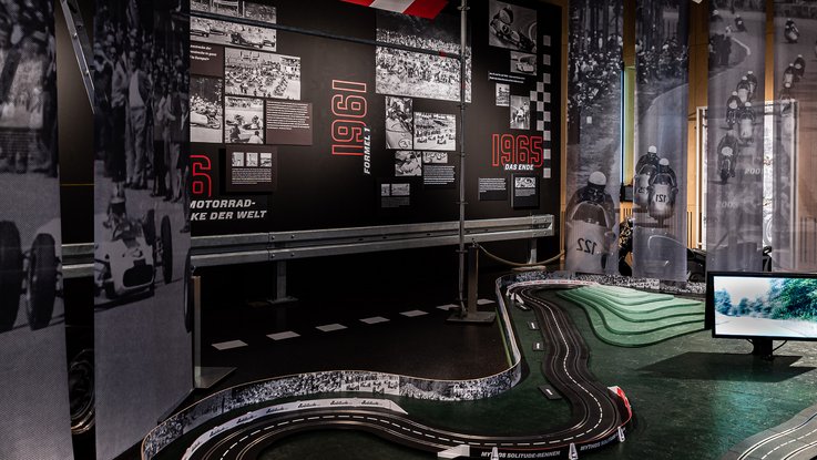 VISUELL Szenografie: Mythos Solitude: Carrerabahn im Mittelpunkt des Bildes und schwarze Informationstafeln im Hintergrund mit schwarz weiß Bildern- Aufschrift in Rot: Formel 1 1961 und das Ende 1965