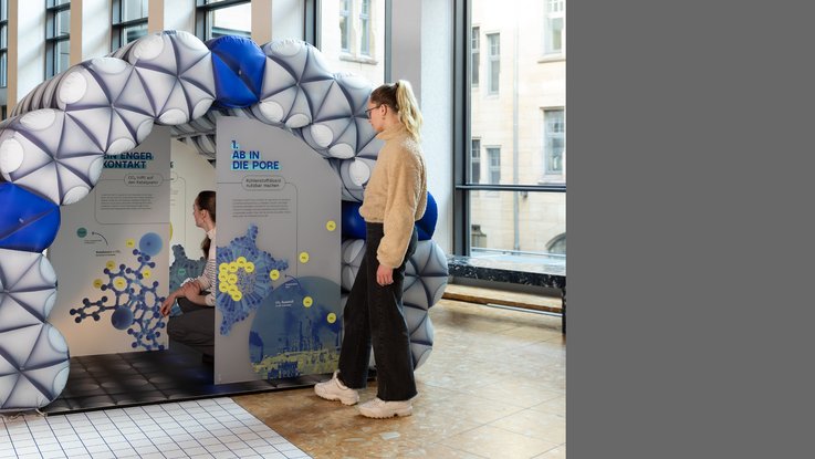 VISUELL Szenografie: Wissenschaftsausstellung „Was kann Katalyse?“ für den Sonderforschungsbereich SFB 1333 der Universität Stuttgart VISUELL Szenografie: Eine Besucherin betrachtet ein begehbares, aufgeblasenes Modell einer Pore in der Wissenschaftsausstellung „Was kann Katalyse?“. Die Struktur des Modells besteht aus Molekülen und ist als Bogen aufgebaut. Im Inneren der Pore befindet sich eine Informationstafel mit dem Titel „Ab in die Pore“. Eine weitere Besucherin befindet sich im Inneren der Pore.