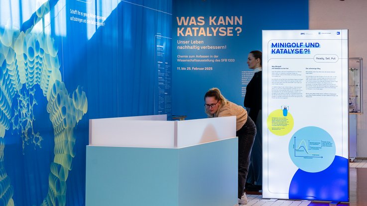 VISUELL Szenografie: Wissenschaftsausstellung „Was kann Katalyse?“ für den Sonderforschungsbereich SFB 1333 der Universität Stuttgart VISUELL Szenografie: Wissenschaftsausstellung: Zwei Besucherinnen stehen neben den Exponaten der Wissenschaftsausstellung „Was kann Katalyse?“. Eine Besucherin bedient das Exponat „Minigolf und Katalyse“ mit einem Schläger, daneben steht eine Informationstafel zu diesem Thema dazu. Die andere Besucherin steht im Hintergrund und betrachtet die blauen Stoffbanner mit weiteren Informationen.