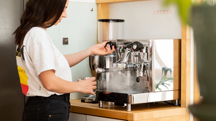 VISUELL Blogbeitrag: Kaffeegenuss: Mitarbeiterin Aline steht an der Kaffee-Ecke bei VISUELL im Studio