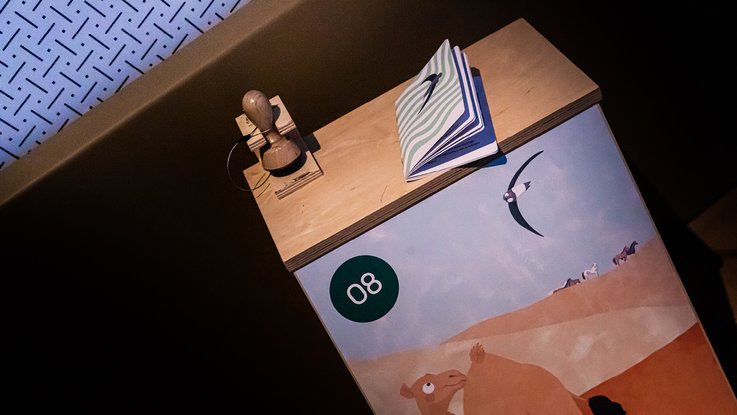 VISUELL Szenografie: Ausstellung „Spurensuche“: Stempelstation mit einem Stempel, einem Stempelbuch und der Illustration des Dromedars