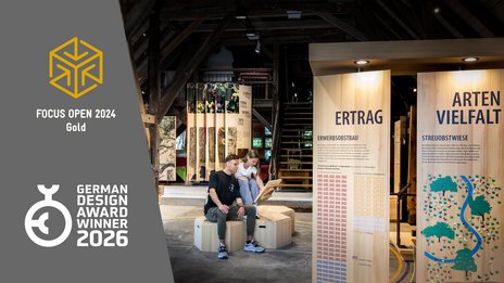 VISUELL Szenografie: Konzeption und Realisierung des Obstbaumuseums in Glems VISUELL Szenografie: Obstbaumuseum: Zwei Besucher sitzen in der Mitte der Ausstellung und betrachten eine Informationstafel in ihren Händen. Links neben dem Bild stehen Logos von Auszeichnungen, die das Projekt erhalten hat: Focus Open 2024 in Gold und German Design Award Winner 2026