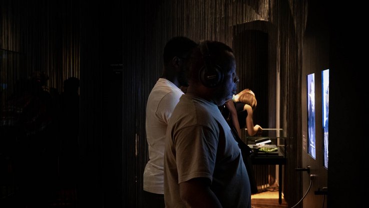 VISUELL Szenografie: Intervention Afrika: Zwei Personen stehen vor einer Medienstation, im Hintergrund stehen zwei Personen über eine Tischvitrine gebeugt.
