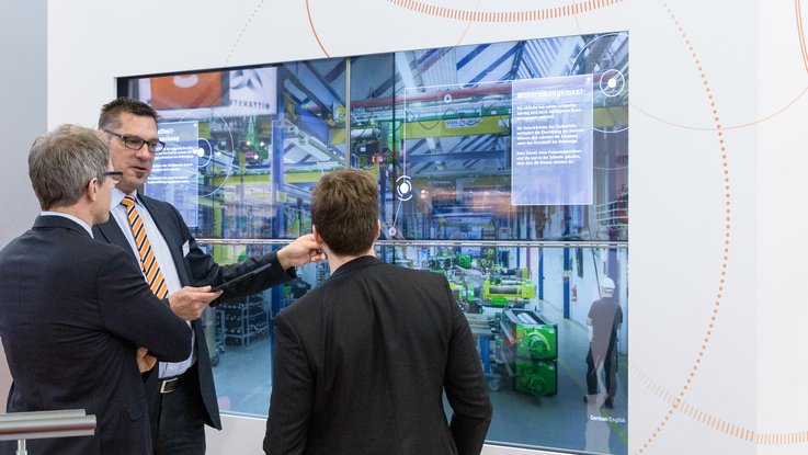 VISUELL Szenografie: Messestand STAHL CraneSystems: Drei Personen bedienen die interaktive Medieninstallation über einen Touchscreen und unterhalten sich