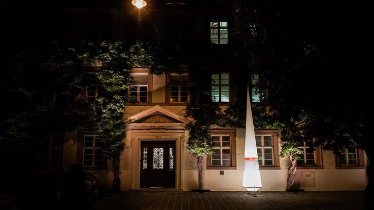 VISUELL Szenografie: Gestaltung und Realisierung des Leitsystems und der Printprodukte für die Nacht des offenen Denkmals in Baden-Baden. VISUELL Szenografie: Nacht des offenen Denkmals: Ein Leuchtkegel mit Banderole steht vor einem Gebäude
