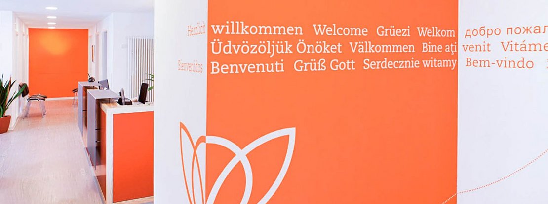 VISUELL Kommunikationsdesign: Corporate Design: „Herzlich willkommen“ in verschiedenen Sprachen zur Begrüßung der Patientinnen und Patienten an der Wand beim Empfang in orange und weiß mit Bildmarke des Logos.