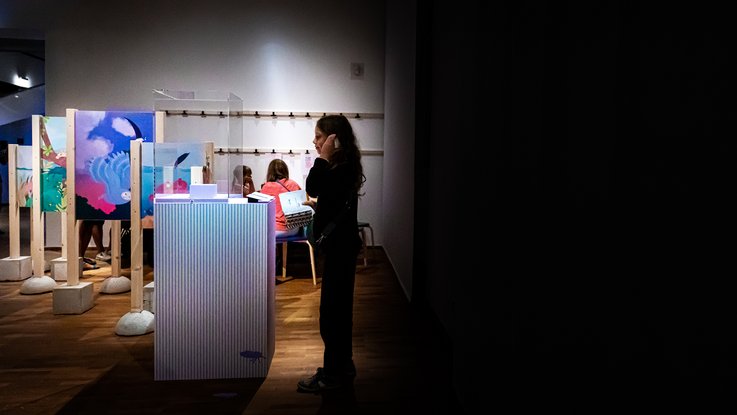VISUELL Szenografie: Ausstellung „Spurensuche“: Besucher steht vor einer Vitrine und hört den Audio-Guide über das Handy