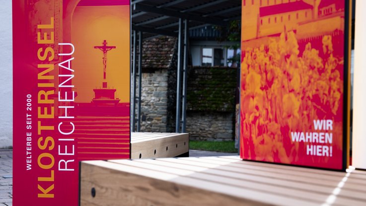 VISUELL Szenografie: Wahre Wunderbänke: Nahaufnahme der Sitzbänke mit Informationseinheiten der Hauptansichtsseite mit plakativen Bildmotiven in magenta und orange mit dem Titel der Welterbestätte sowie der Aufschrift WIR WAHREN HIER!