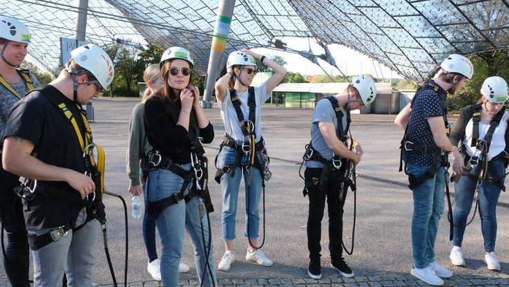 VISUELL Blogbeitrag: Weiterbildung in Innsbruck und München: Das VISUELL-Team steht mit Helm und Gurt vor dem Olympiastadion in München