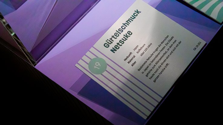 VISUELL Szenografie: Ausstellung „Spurensuche“: Objektschild eines Exponats mit lilalen Streifen am Rand - Aufschrift: Gürtelschmuck Netsuke