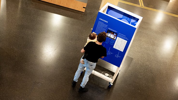 VISUELL Szenografie: Jubiläumsausstellung: Ein Mann mit einem Kind auf dem Arm steht vor einem hohen Rollmöbel und schaut das Video, er trägt Kopfhörer
