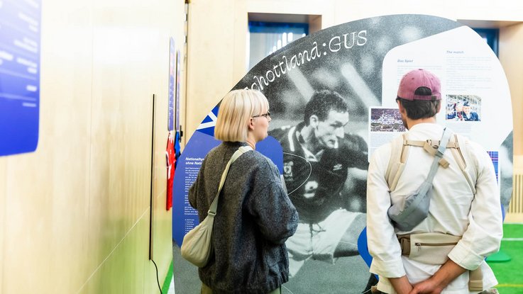 VISUELL Szenografie: EURO Legends Stuttgart: Zwei Personen stehen vor dem Aufsteller über die schottische Mannschaft und lesen die Texte und schauen die Fotos an.