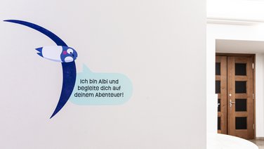 VISUELL Szenografie: Ausstellung „Spurensuche“: Wandbeschriftung mit einer Illustration von Albi, dem Mauersegler - Aufschrift: Ich bin Albi und begleite dich auf deinem Abenteuer!