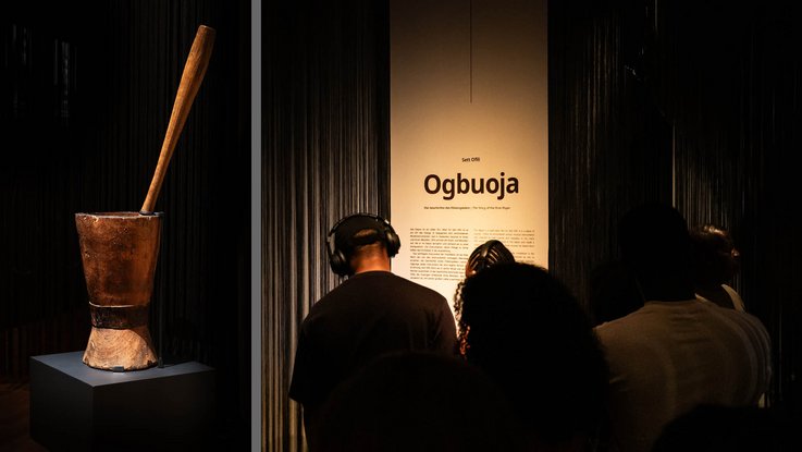 VISUELL Szenografie: Intervention Afrika: Links ist ein Mörser mit einem Stößel auf einem Podest zu sehen, rechts stehen Personen mit Kopfhörern vor einem Banner mit Schrift.