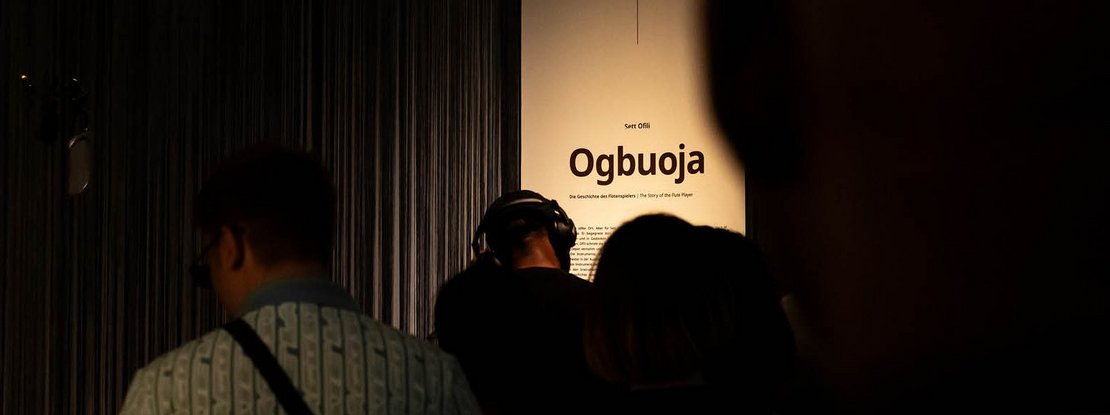 VISUELL Szenografie: Intervention Afrika: Viele Menschen stehen vor einem Banner mit dem Titel der Intervention von Sett Ofili „Ogbuoja: The Story of a Flute Player“