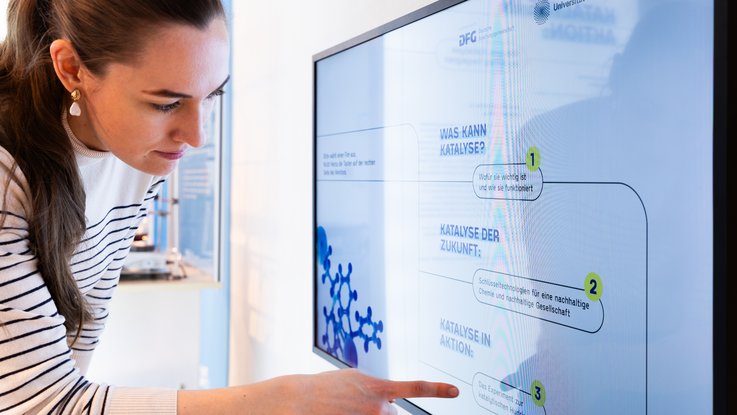 VISUELL Szenografie: Wissenschaftsausstellung „Was kann Katalyse?“ für den Sonderforschungsbereich SFB 1333 der Universität Stuttgart VISUELL Szenografie: Wissenschaftsausstellung: Nahaufnahme einer Besucherin die vor einem Bildschirm steht und auf Elemente der dort abgespielten Informationsgrafik zeigt.