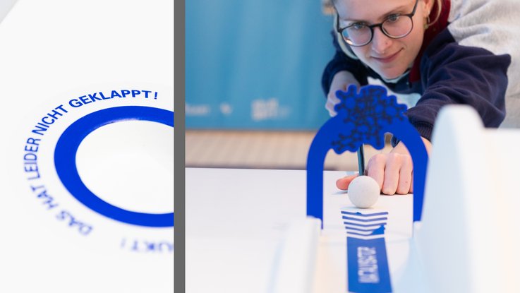 VISUELL Szenografie: Wissenschaftsausstellung „Was kann Katalyse?“ für den Sonderforschungsbereich SFB 1333 der Universität Stuttgart VISUELL Szenografie: Wissenschaftsausstellung: Links: Detailaufnahme einer blauen Beklebung des Exponats „Minigolf und Katalyse“. Rechts: Frontale Nahaufnahme einer Besucherin die dieses Exponat mit einem Schäger bedient und durch einen blauen Bogen zielt.