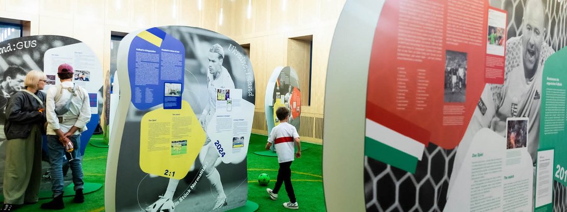 VISUELL Szenografie: EURO Legends Stuttgart: Eine Frau und ein Mann stehen zwischen überlebensgroßen Aufstellern von Fußballspielern, ein Junge dribbelt mit einem Ball durch den Parcours.