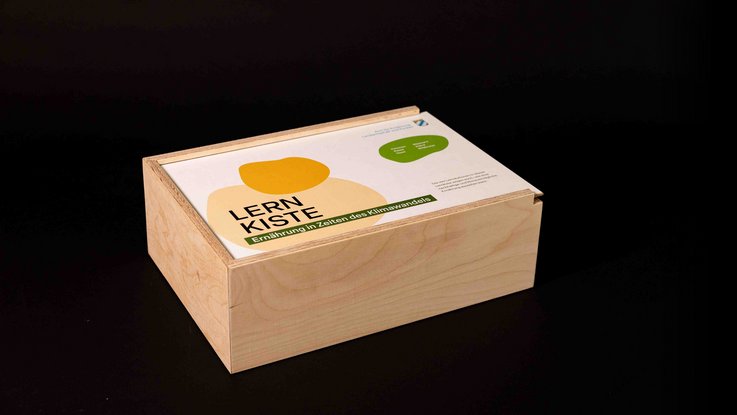VISUELL Szenografie: Pädagogische Lernkiste: Geschlossenen Holzkiste mit grün und gelben organischen Elemente- Aufschrift: Lernkiste, Ernährung in Zeiten des Klimawandels