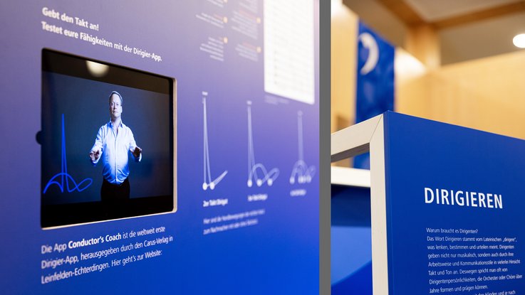 VISUELL Szenografie: Jubiläumsausstellung: links Nahaufnahme des ins Möbel eingelassenen Tablets mit Dirigier-App Conductor’s Coach, rechts Nahaufnahme des niedrigen Rollmöbels mit der Überschrift Dirigieren sowie Text
