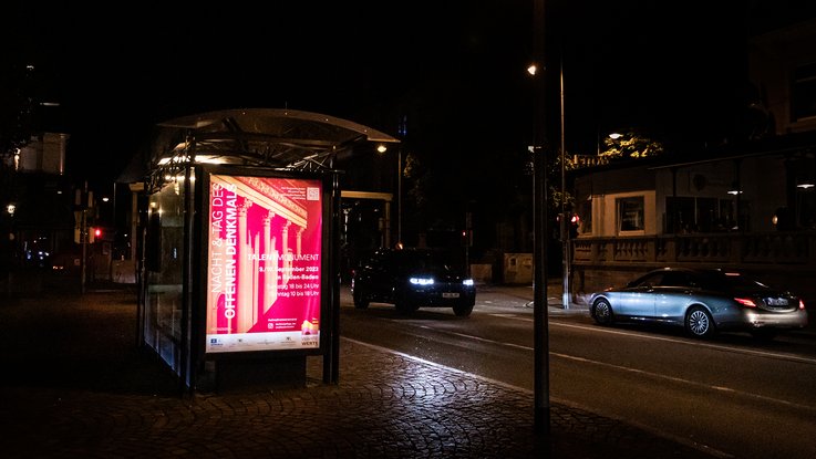 VISUELL Szenografie: Gestaltung und Realisierung des Leitsystems und der Printprodukte für die Nacht des offenen Denkmals in Baden-Baden. VISUELL Szenografie: Nacht des offenen Denkmals: Das Plakat der Nacht des offenen Denkmals hängt bei Nacht an einer Bushaltestelle