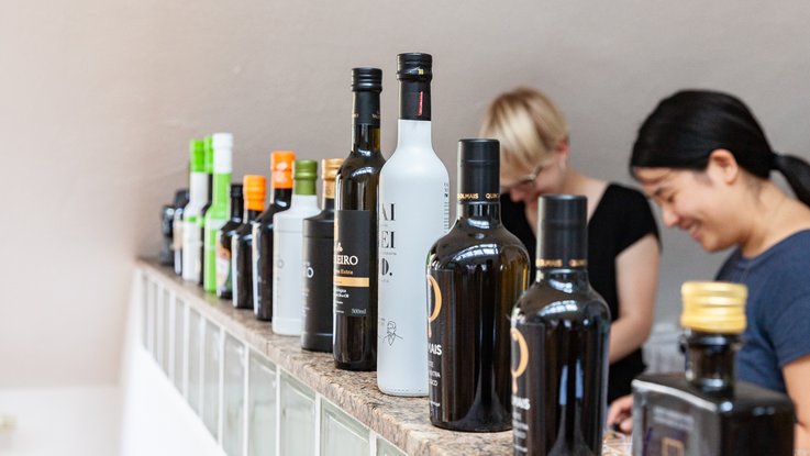 VISUELL Blogbeitrag: Das Event unterm Dach mit dem Thema „Taste the best“ – das Olivenöl-tasting. VISUELL Blogbeitrag: Event unterm Dach– das Olivenöl-tasting: Eine Reihe verschiedener Olivenölflaschen steht auf einer Theke