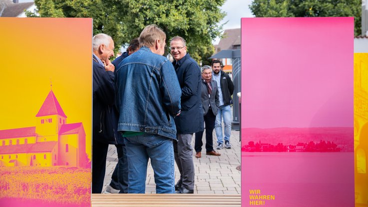 VISUELL Szenografie: Informationseinheiten für die sieben UNESCO-Welterbestätten in Baden-Württemberg in Zusammenarbeit mit dem Ministerium für Landesentwicklung und Wohnen Baden-Württemberg VISUELL Szenografie: Wahre Wunderbänke: Eine Gruppe von Besuchenden steht vor einer Sitzbank, die Rückseite zweier Informationstafeln mit Bildmotiven in magenta und gelb ist sichtbar